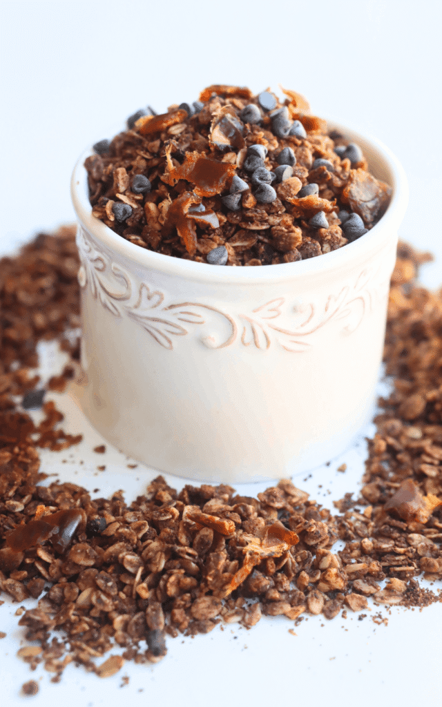 vertical photo showing a white bowl with healthy homemade chocolate granola inside and around on a white surface