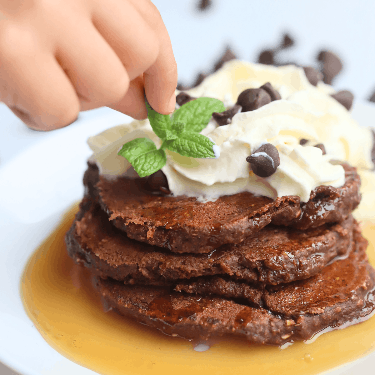 a hand placing a mint leaf on top of a pile of chocolate pancakes topped with homemade whipped cream