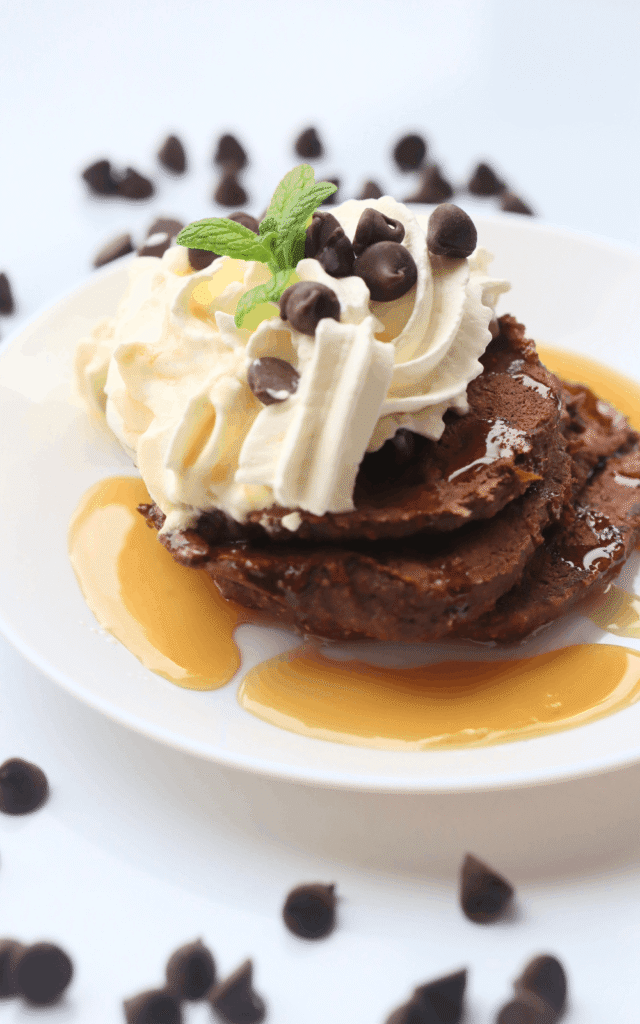 a pile of chocolate protein pancakes on a white plate and topped with whipped cream and maple syrup and a mint leaf