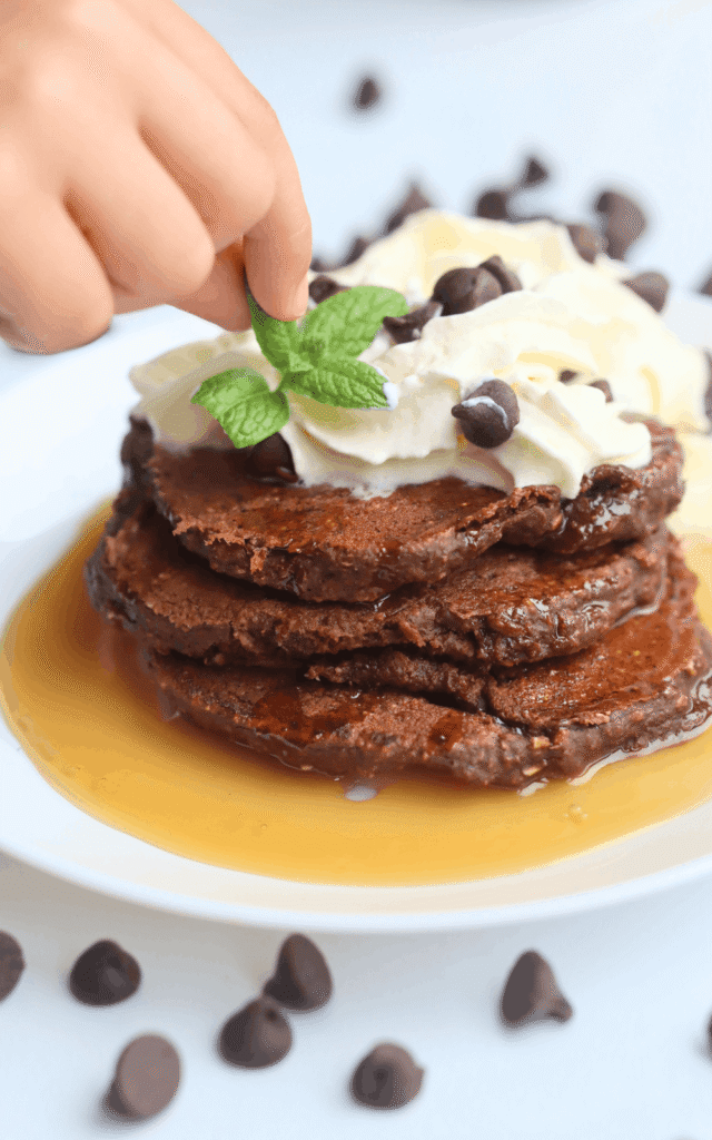 a hand placing a mint leaf on top of a pile of chocolate pancakes topped with homemade whipped cream