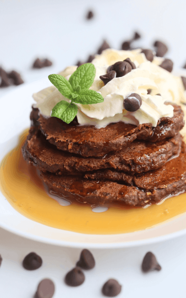 a pile of chocolate protein pancakes on a white plate and topped with whipped cream and maple syrup and a mint leaf