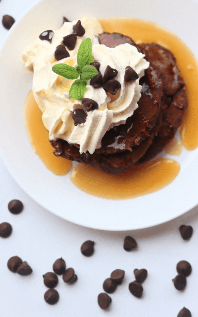 top-down view of a pile of chocolate protein pancakes on a white plate and topped with whipped cream and maple syrup and a mint leaf