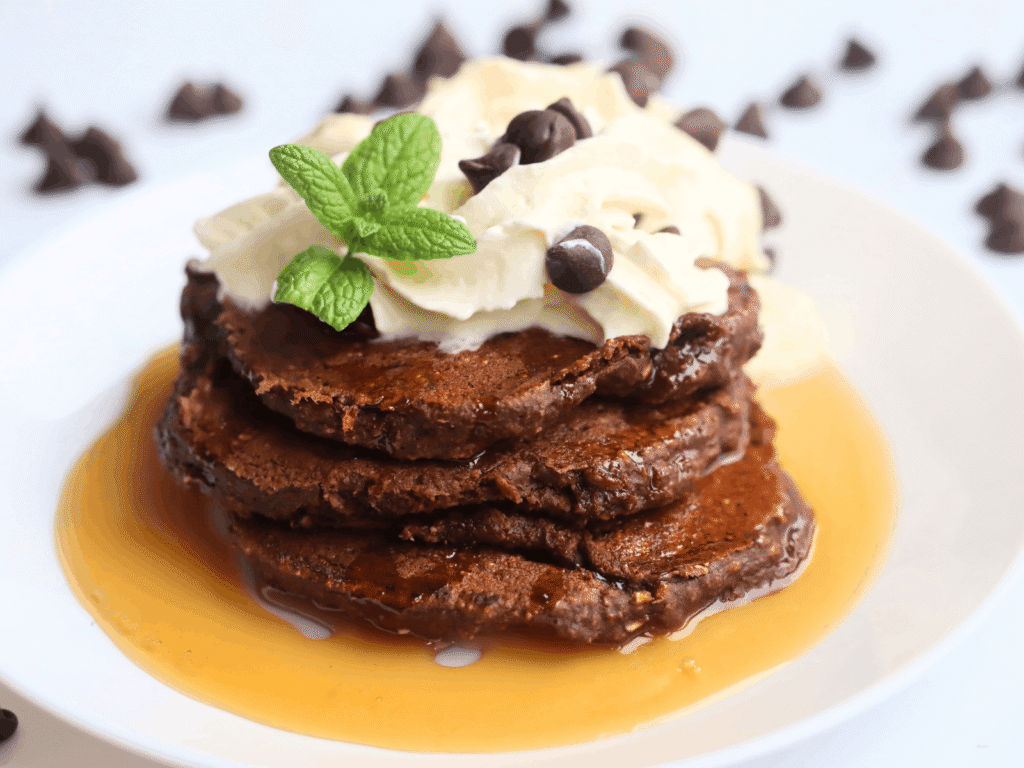 a pile of chocolate protein pancakes on a white plate and topped with whipped cream and maple syrup and a mint leaf